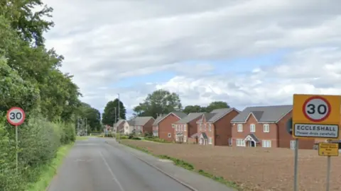 Google Tree line one side of the road with new homes on the other. Two 30mph signs can be seen with one naming Eccleshall