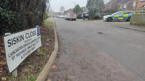 A police vehicle outside a property in Siskin Close. There are a number of properties and trees nearby, and police tape in front of the house. 