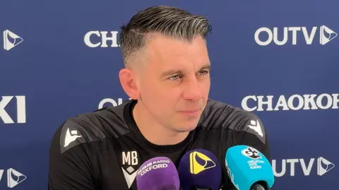 Matt Bloomfield has short dark hair slicked over to one side and is wearing an Oxford United shirt. He is sitting in front of a collection of microphones with a blue and white sponsors board behind him.