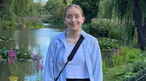 Libby smiling into the camera in front of a pond with lily pads, plants and flowers. She has blonde hair which is tied back and is wearing a blue and white striped shirt with a white tank top underneath. She is also wearing a black cross-body bag and a simple silver necklace which is silver with a small pendant. 