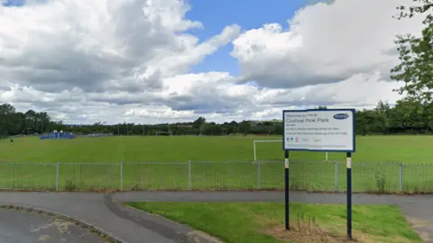 A sign reading Welcome to Coxhoe Park in front of a large green field. There are football goals on the field which is bordered on three sides by trees.