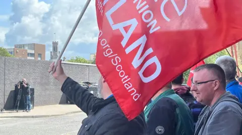 People protesting outside a Stagecoach depot with a Unite union red banner. There is a photographer taking pictures across the street 