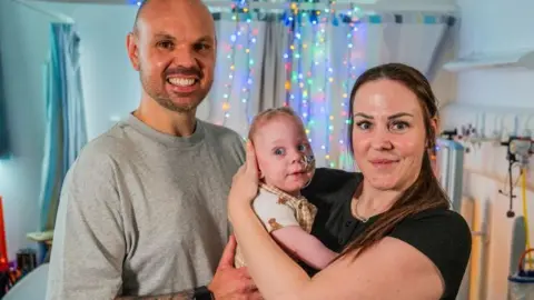 Chris Watt Photography Kirsty is holing her son, Kobi, in her arms. She hass long brown hair. Daniel is standing beside them and smiling. He has a shaved head and stubble on his chin. He is wearing a grey top.