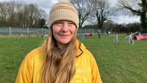A woman with long, light brown hair, a beige striped woolly hat and a yellow waterproof jacket stands in a dog agility field. In the background, tunnels, jumps and other obstacles can be seen. 