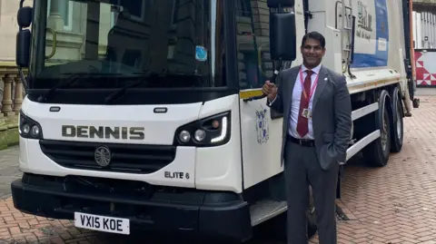 A man standing next to a bin lorry on red brick road. 