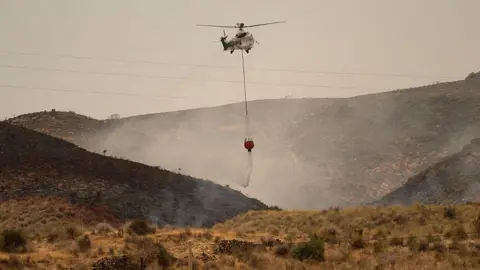 Getty Images A wide angle photo showing a helicopter dropping water on a grass and shrub wildfire in Spain.