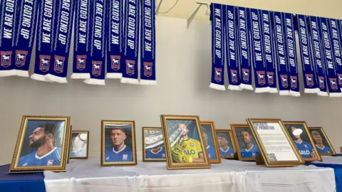 Ipswich Central A table where artistic portraits of football players have been placed in small frames. Matching blue football scarfs with the words "we are going up" hang above from a wall in a line.