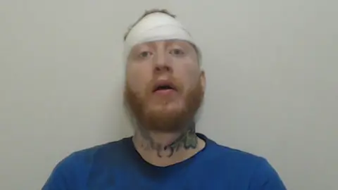 A mugshot of Stephen Ball, wearing a head bandage. He has a ginger beard, tattoos on his neck, and is wearing a blue t-shirt.