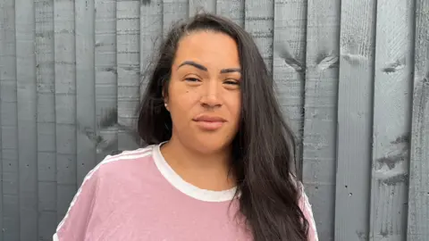 Tony Fisher/BBC Sasha Field with black long hair looking at the camera. She is in front of a wooden fence and is wearing a pink top.