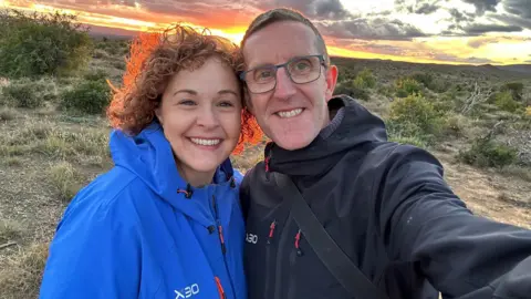 Amy Attwood A smiling couple take a selfie outdoors in a scrubland landscape at sunset. The woman on the left has curly red hair and wears a bright blue jacket. The man on the right wears glasses, a black jacket and is holding the camera. Dramatic orange and grey clouds fill the sky behind them.