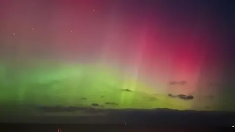 BBC Weather Watchers/Sarah n Essie A close-up of the sky which has a few wispy clouds. The Northern Lights are shining green and pink.