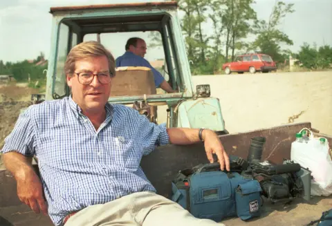 Stourton sits on the back of a tractor, being pulled along in a trailer, wearing a shirt and glasses while sitting next to camera equipment and bottled water, with a red car sitting on a dusty tree-lined road in the background.