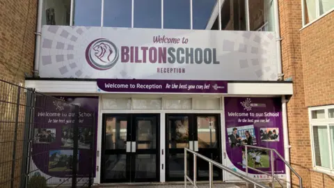 The entrance to a school building. Above two set of black double doors is a white sign that says welcome to Bilton School in purple and grey text. On either side of the doors are graphics including pictures of children and the words "welcome to our school, be the best you can be!"
