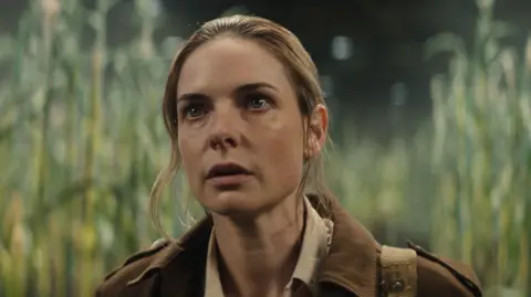 Rebecca Ferguson acting as Jules in Silo, stares up at something. She is surrounded by a field of corn and wears a brown shirt and has her hair scraped back.