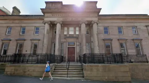 Google A two-storey stone building with steps leading up to an entrance which has four pillars and engraved test reads Liverpool Institute and school of art 1825. A woman with long brown hair and wearing a white t-shirt black shorts and white trainers and carrying a blue bag walks past.