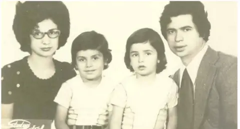 Acun Ilicari A sepia toned formal family photograph. A woman with a large 1960s hair style and thick framed glasses is to the left of the picture, she is wearing a dark scalloped neck top. short-sleeved top. Two children wearing identical clothes sit between her and a man in a suit, shirt and tie, he has dark wavy hair.