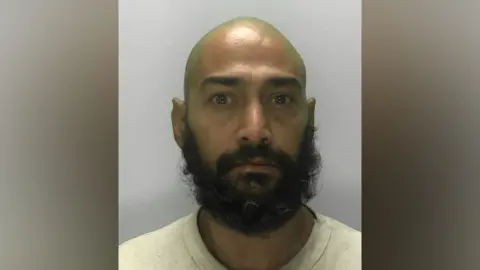 Afzul Veshima, a man with a bald head and bushy black beard, stares straight ahead in police photo 