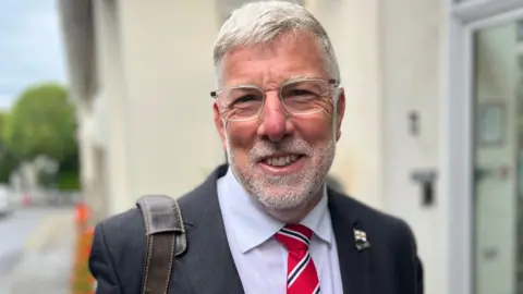 Deputy Adrian Gabriel, a man with grey hair, a grey beard and glasses. He's wearing a black suit with a Guernsey flag lapel badge. He has a bag strap on his shoulder. 