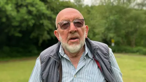 Man with white beard and dark glasses, wearing a blue and white striped shirt and gilet, posing for a photo with blurred trees and parkland behind him