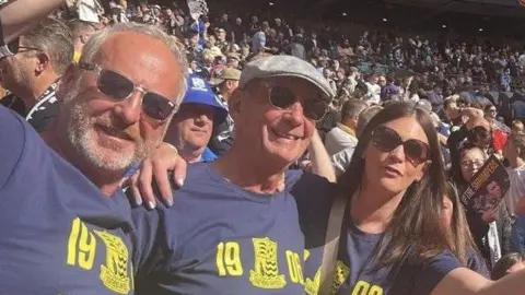 Julian Wantling with a man and a woman inside Wembley in 2025. They are wearing blue Southend United T-shirts and sunglasses.