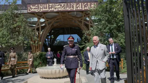 PA Media King Charles III and someone in military uniform are walking towards a gate which is in front of a large mosque which has scripture above its entrance.