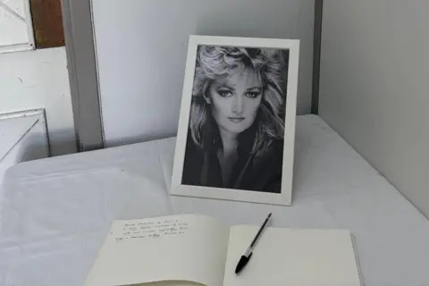 A black and white picture of Bonnie Tyler behind a book with a pen. A message can be seen written on the left hand page. 