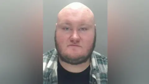 Mugshot of Andrew Weir. He is a large man with a bald head and dark stubbly beard around his chin and sideburns. He is wearing a green and white check shirt over a black t-shirt.