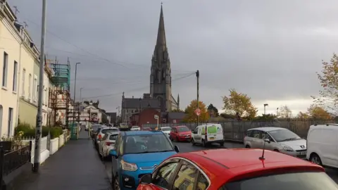 BBC A row of terraced townhouses with a long line of parked cars outside each of them. There is a long line of cars parked on the other side of the street also. A cathedral can be seen in the background