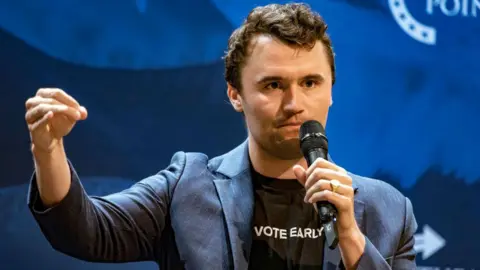 Getty Images Charlie Kirk at an event with a microphone. His shirt says 'vote early'