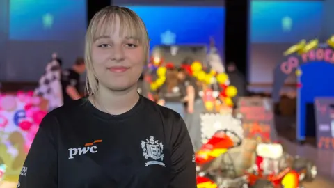 Maja is wearing a black PwC T‑shirt stands in front of a decorated go‑kart display on a stage with blue lighting and event signage in the background