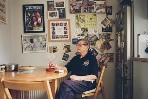 Craig Mcintosh Moira sitting at the table of her kitchen, the wall behind her is covered with framed photographs.
