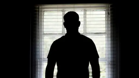 The silhouette of a man from the back looking out of a bay window with a slatted blind