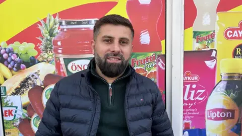 Kate Bradbrook/BBC Shahrim Ali with short dark hair and medium-length beard, wearing a dark top and blue gilet, standing in front of a shop window which has illustrations of various products.