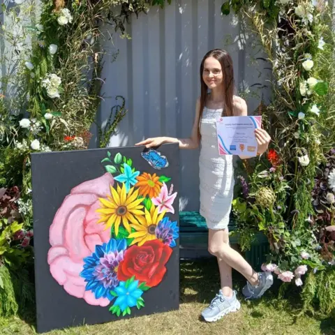 Victoria Glover Freya is wearing a white dress and has long, straight brown hair. Freya is standing next to a colourful piece of artwork. The painting is of part of a brain on the left side and vibrant flowers on the right side. Freya is holding a certificate as this was the artwork that won the prize at the Llangollen Eistedfodd.