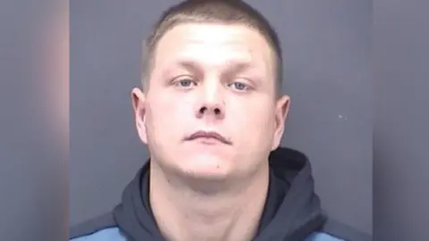 A mugshot of a man with short light brown hair. He is wearing a blue hoodie.