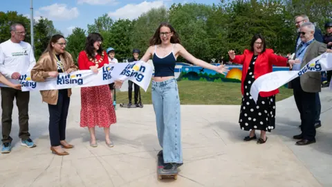 CGF Photography A young woman wearing jeans and a blue top skateboarding through a ribbon which has "Luton Rising" on it and is being held by men and women. She is in a skatepark with trees in the background.