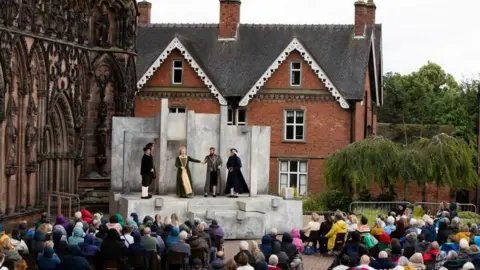 Lichfield Festival Dozens of people sit outside a cathedral watching a theatre performance on an outdoor stage. The cast can be seen wearing costumes. A cathedral building and a house can be seen in the background.