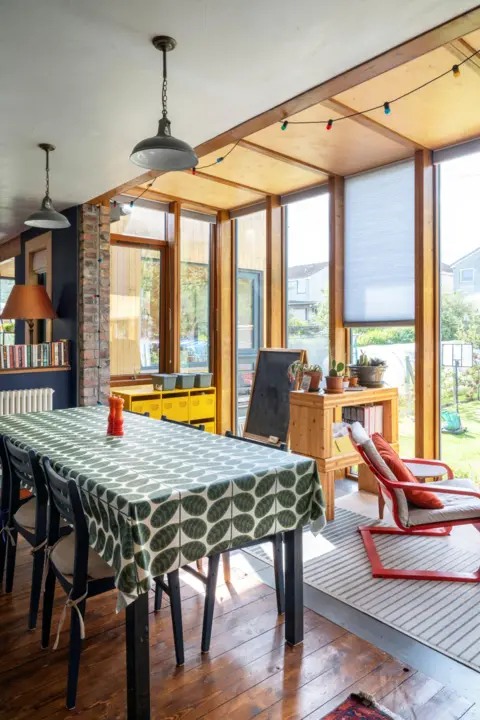 IWC Media A bright dining area sits within a sunlit extension, featuring large wooden-framed windows that look out onto a green garden.
A rectangular table covered with a patterned cloth is surrounded by dark chairs, with pendant lights and colorful string lights hanging above.
A mix of natural wood, exposed brick, houseplants, and shelving adds warmth and a relaxed, lived-in feel to the space.