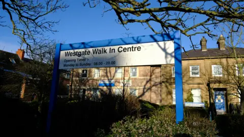 BBC A blue and white sign reading: "Westgate Walk In Centre" sits in front of two, two-storey buildings.