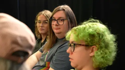 Richard Knights/BBC An candid image showing three people in a focus group listening while another person off-camera is speaking. Benji Rayson is in the middle of the image, looking towards the left of the frame. They are wearing a grey t-shirt and have straight brown hair that hangs below their shoulders. They have dark-rimmed rectangular glasses and have a neutral expression on their face.