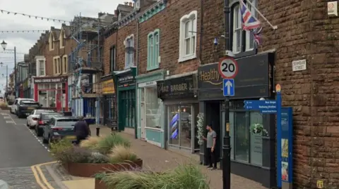 A view of the shops on Senhouse Street in Maryport. A range of shops can be seen and planters with different types of grass growing in them can be seen on the pavements in front of the shops.