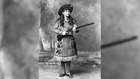 Getty Images Oakley, shown with long brown curly hair beneath a wide-brimmed hat, is posing with a rifle