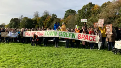 Martin Heath/BBC A crowd of people standing in a grass park with trees behind, some holding banners saying things like "keep off the grass".  A number of them are holding up a banner saying "Saving our town green space"