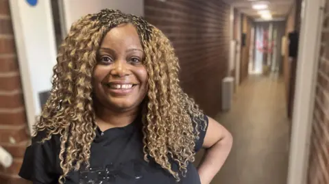 A woman with curly hair, with a beaming smile for the camera and wearing a short-sleeved black T-shirt