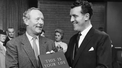 A black-and-white mid-shot of two men, half-turned towards one another and smiling on a television studio set with nondescript plain backdrops. The man on the right is in his late 30s, with slightly curly black hair, a black jacket, a white shirt and dark tie. He is a few inches taller than the man on the left, who is in his early 50s with receding grey hair, a grey pinstriped jacket, white shirt and pale-shaded tie. The man on the right is handing the man on the left a large hardback book with ‘This is Your Life’ written on it in block capitals. Behind the two men can be seen various members of a studio audience, or possibly invited guests for the television programme.
