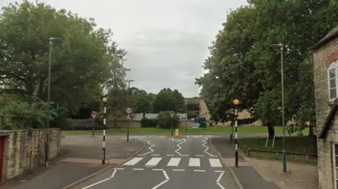 A Google streetview of Querns Lane, Cirencester, showing a zebra crossing and trees either side.
