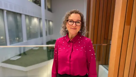 Woman in pink blouse with short curly hair and glasses stood in hallway of council house