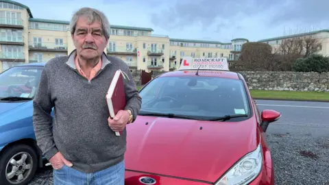 Wayne Stokes pictured with short grey hair and a moustache. He is wearing a grey quarter-zip knitted jumper which has a salmon pink collar poking out the top. He has his hand tucked into the pocket of his blue denim jeans and is clutching a burgundy notebook in his other hand. Wayne stands in front of a burgundy Ford Fiesta which has the name of his driving school 'Roadcraft' on the top with a red 'L'