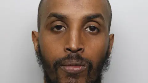 A mugshot of a bald man with a thin mid-length black beard. He has dark eyes and arched eyebrows. He has a neutral expression.