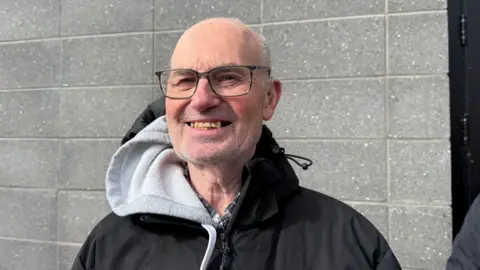 A man with grey stubble, a bald head and glasses smiles into the camera. He is wearing a black jacket with a grey hood and is standing in front of a grey brick wall.